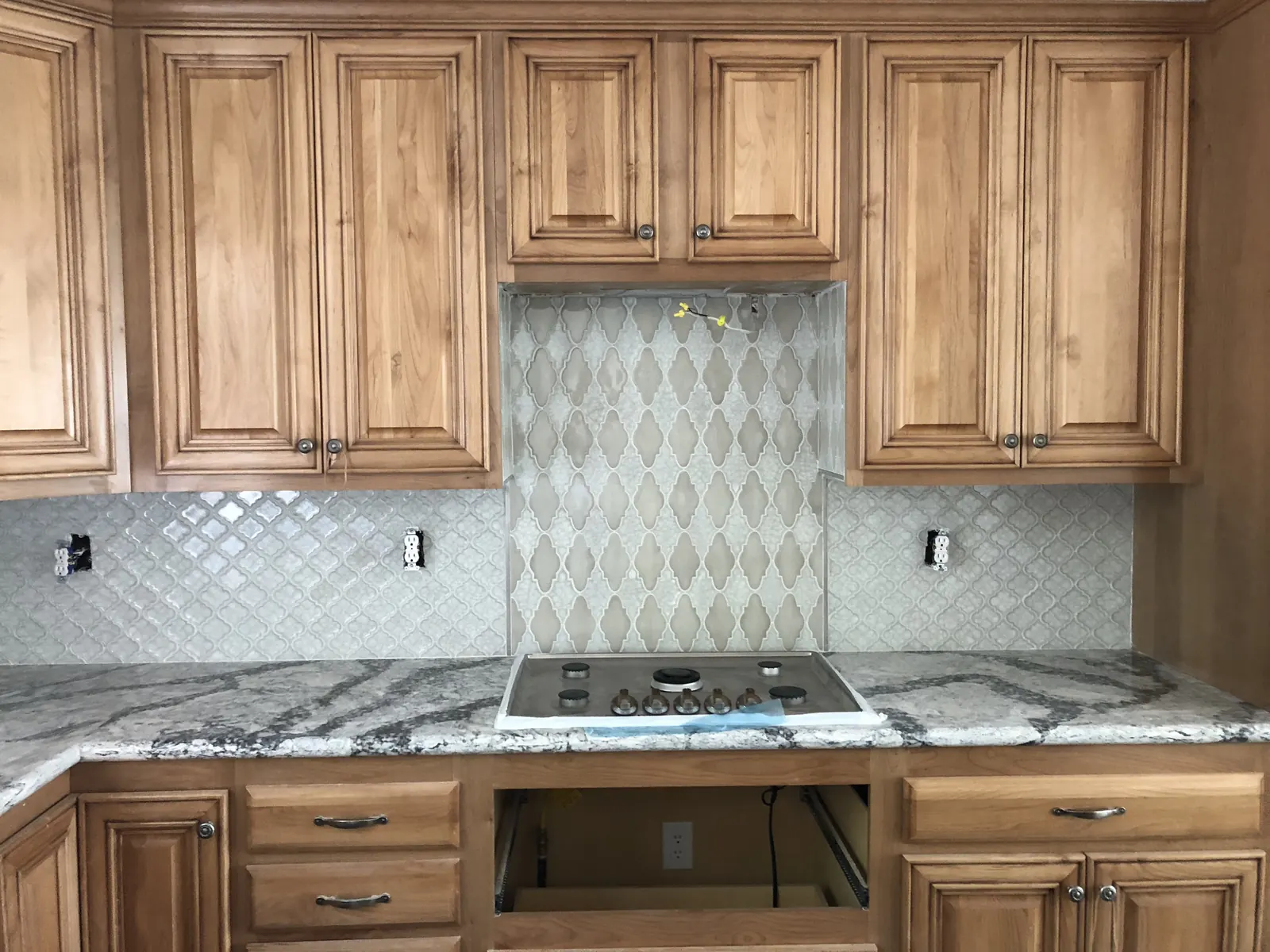 Gray patterned backsplash with a taller decorative tile panel above a cooktop and cuts around outlets