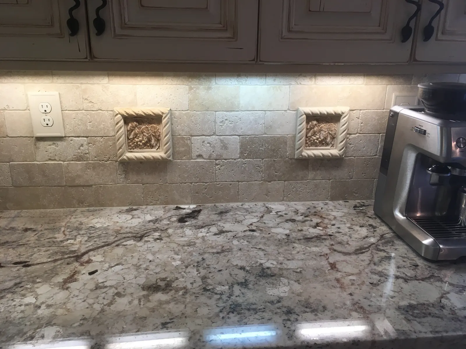 Textured beige kitchen backsplash with framed decorative accents, an outlet, and under-cabinet lighting